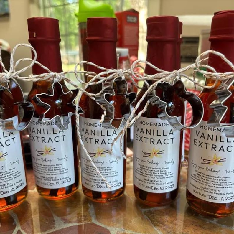 May include: Five small glass bottles filled with brown liquid and labeled "Homemade Vanilla Extract". Each bottle is decorated with a white ribbon and a metal cookie cutter in the shape of a star. The bottles are arranged in a row on a brown surface.