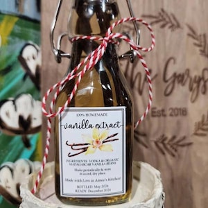 May include: A small glass bottle of homemade vanilla extract, labeled with ingredients and instructions. The label reads "100% Homemade vanilla extract" and includes the text "Made with Love in Aimee's Kitchen!" The bottle is tied with red and white twine.