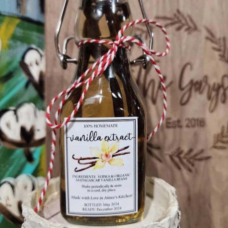 May include: A small glass bottle of homemade vanilla extract, labeled with ingredients and instructions. The label reads "100% Homemade vanilla extract" and includes the text "Made with Love in Aimee's Kitchen!" The bottle is tied with red and white twine.