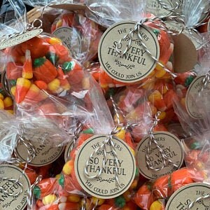 May include: A close-up of small bags filled with orange and yellow candy corn. Each bag has a brown paper circle tag with the text "The [blank]ers So Very Thankful You Could Join Us".