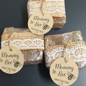 May include: Three clear, honey-colored soap bars wrapped in burlap and lace with a brown circle tag that reads "Mommy to Bee" with a bee illustration and the text "EMMA & BABY PHOEBE 2.1.20".