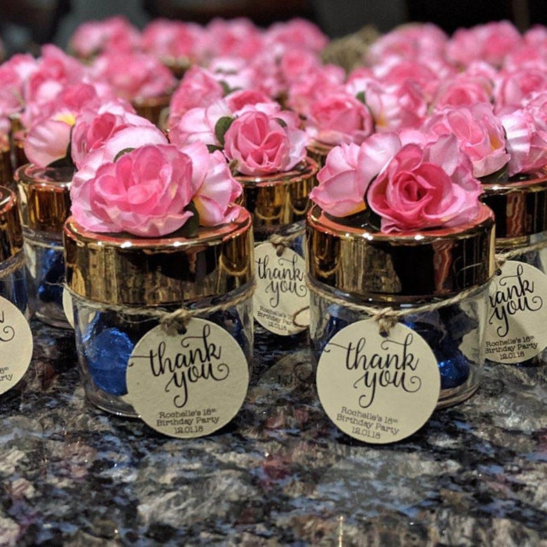 May include: Small glass jars with gold lids decorated with pink flowers and a round thank you tag. The tag says "Thank You" with the text "Rochelle's 18th Birthday Party 18.01.18".