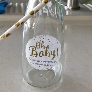 Op de afbeelding: Een heldere glazen fles met een wit deksel en een gouden rietje. De fles heeft een wit en gouden sticker met de tekst "Oh Baby!" met de tekst "Ty & Nicole's Baby Shower September 29, 2018."