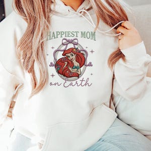 May include: Cream-colored hoodie with the text "Happiest Mom on Earth" in green and purple. The design features a cartoon illustration of a mermaid with red hair and a green top, surrounded by a decorative frame.