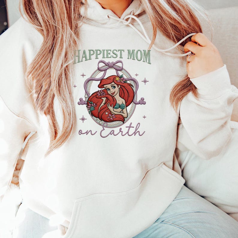 May include: Cream-colored hoodie with the text "Happiest Mom on Earth" in green and purple. The design features a cartoon illustration of a mermaid with red hair and a green top, surrounded by a decorative frame.