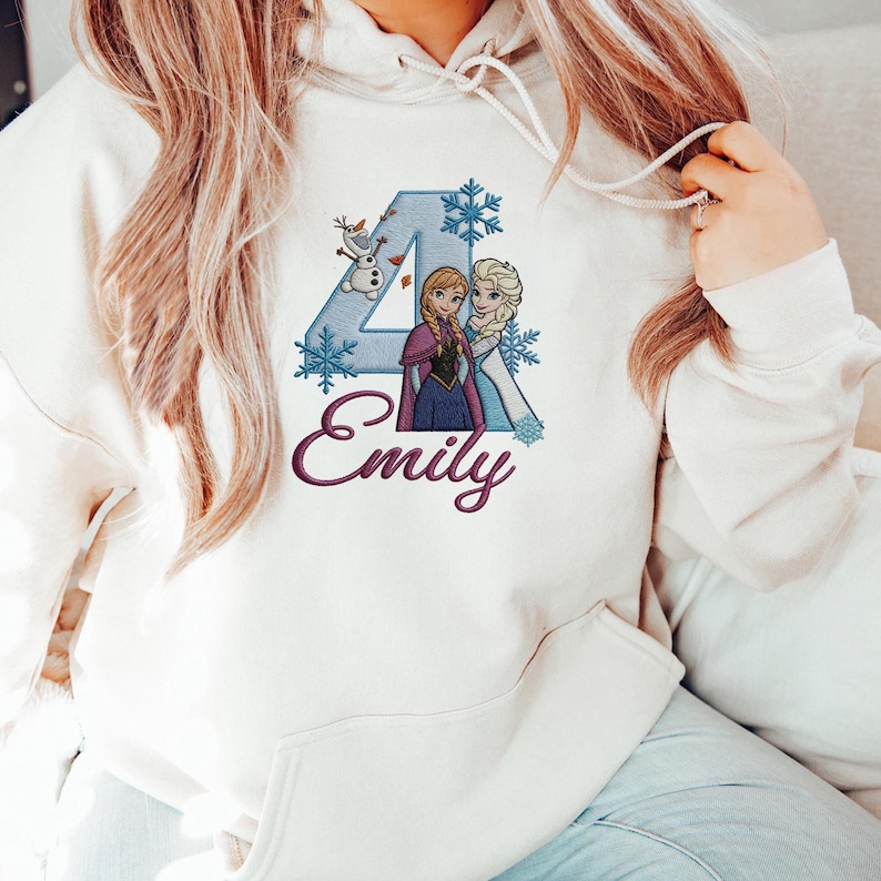 Puede incluir: Sudadera con capucha de color crema con un dise&ntilde;o bordado colorido. El dise&ntilde;o incluye el n&uacute;mero 4, copos de nieve y dos personajes de dibujos animados. El nombre "Emily" est&aacute; bordado en cursiva debajo del dise&ntilde;o.