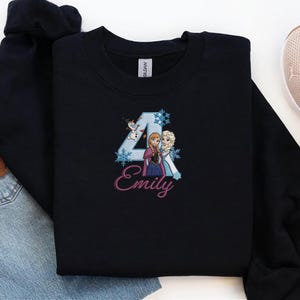 Puede incluir: Una sudadera negra con un dise&ntilde;o bordado. El dise&ntilde;o incluye el n&uacute;mero 4, copos de nieve y los personajes Anna, Elsa y Olaf de la pel&iacute;cula Frozen. El nombre "Emily" est&aacute; escrito en rosa.