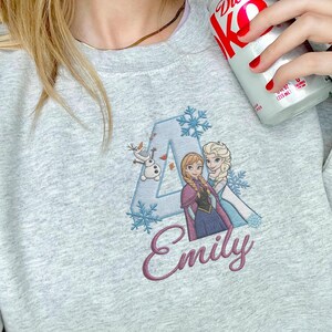 Puede incluir: Sudadera gris claro con un dise&ntilde;o bordado. El dise&ntilde;o incluye la letra "A", copos de nieve y los personajes Anna, Elsa y Olaf de la pel&iacute;cula Frozen. El nombre "Emily" est&aacute; escrito en cursiva debajo.