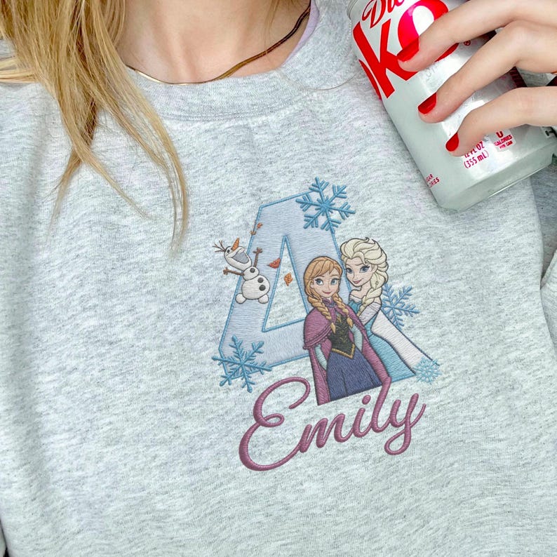 Puede incluir: Sudadera gris claro con un dise&ntilde;o bordado. El dise&ntilde;o incluye la letra "A", copos de nieve y los personajes Anna, Elsa y Olaf de la pel&iacute;cula Frozen. El nombre "Emily" est&aacute; escrito en cursiva debajo.