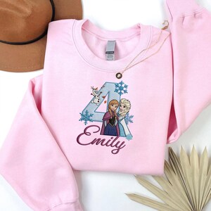 Puede incluir: Sudadera rosa claro con un gran n&uacute;mero 4 bordado con un dise&ntilde;o de copo de nieve azul, con los personajes Anna y Elsa de la pel&iacute;cula Frozen. El nombre "Emily" est&aacute; bordado en cursiva debajo.