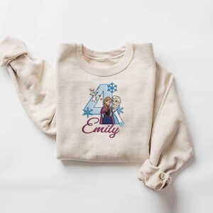 Puede incluir: Una sudadera color crema con un gr&aacute;fico del n&uacute;mero 4, dos personajes de dibujos animados, copos de nieve y el nombre "Emily" en cursiva. El dise&ntilde;o es en tonos azules, morados y blancos.