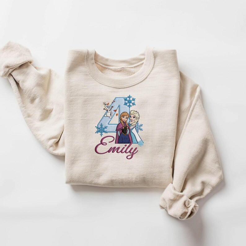 Puede incluir: Una sudadera color crema con un gr&aacute;fico del n&uacute;mero 4, dos personajes de dibujos animados, copos de nieve y el nombre "Emily" en cursiva. El dise&ntilde;o es en tonos azules, morados y blancos.