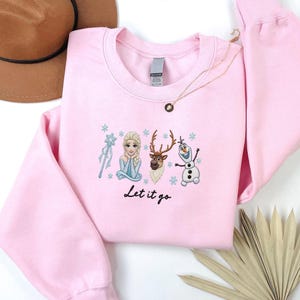 May include: A light pink sweatshirt featuring embroidered characters from a popular animated film. The design includes a princess, a reindeer, and a snowman, with the phrase "Let it go" below. A gold necklace and a brown hat are also visible.