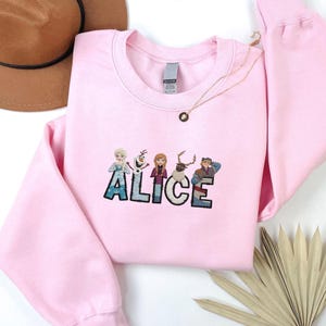 Może przedstawiać: Jasnor&oacute;żowa bluza z wyszytym imieniem "ALICE" w kolorowych literach i postaciami z kresk&oacute;wek. Złoty naszyjnik z wisiorkiem jest zarzucony na bluzę. Brązowy kapelusz jest widoczny w lewym g&oacute;rnym rogu.