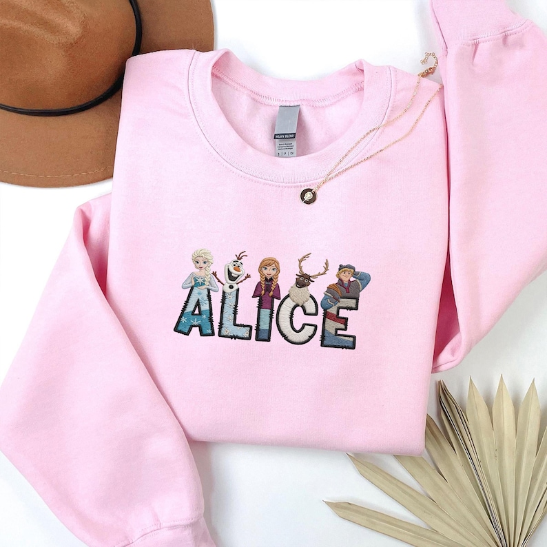 Może przedstawiać: Jasnor&oacute;żowa bluza z wyszytym imieniem "ALICE" w kolorowych literach i postaciami z kresk&oacute;wek. Złoty naszyjnik z wisiorkiem jest zarzucony na bluzę. Brązowy kapelusz jest widoczny w lewym g&oacute;rnym rogu.