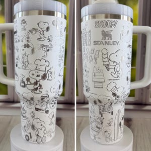 May include: White insulated tumbler with a handle, featuring a repeating pattern of Snoopy characters in various poses. The tumbler has a white lid and handle, and the text "SNOOPY" and "STANLEY" are visible.