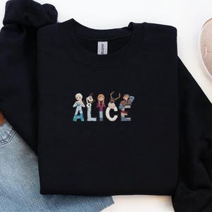 Może przedstawiać: Czarna bluza z okrągłym dekoltem z wyszytym imieniem "ALICE" w kolorowych literach, z postaciami z kresk&oacute;wek. Bluza wykonana jest z miękkiego materiału i ma klasyczny kr&oacute;j. Swobodny i wygodny element garderoby do noszenia na co dzień.