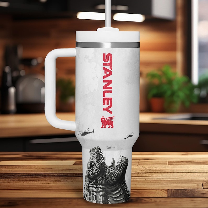 K&ouml;nnte beinhalten: Wei&szlig;er isolierter Becher mit Henkel und Strohhalm. Der Becher zeigt den roten Text "STANLEY" und eine Grafik eines ge&ouml;ffneten Krokodilmauls. Kleine Hubschraubergrafiken sind ebenfalls vorhanden.