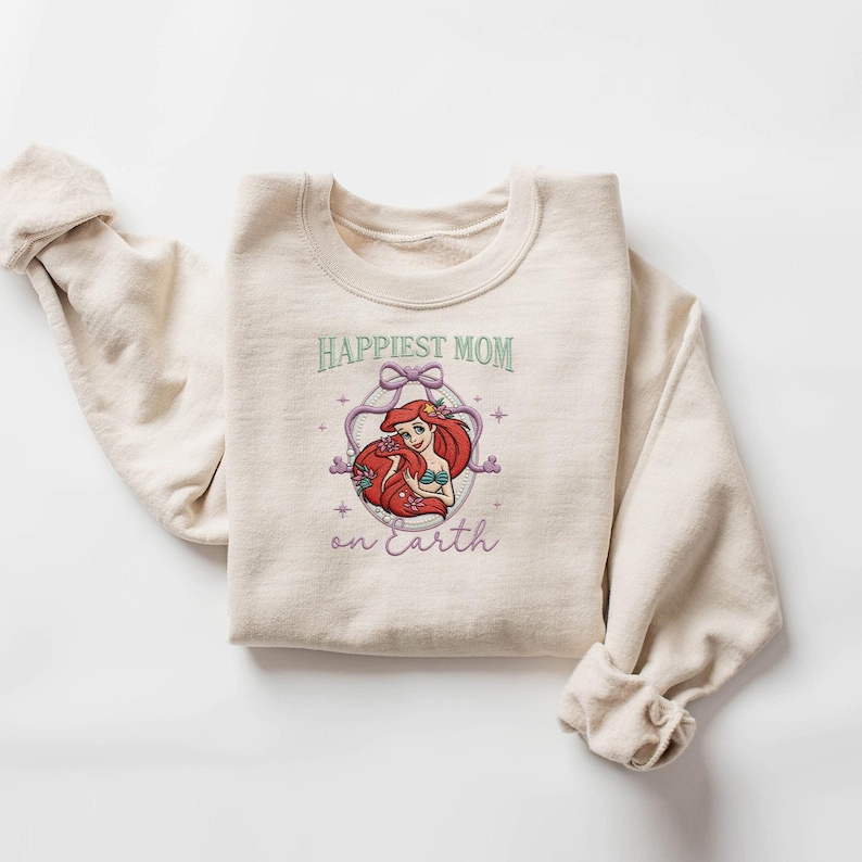 May include: A beige sweatshirt with the text "Happiest Mom on Earth" in green and purple. The graphic features a cartoon illustration of a red-haired mermaid inside a decorative frame. The sweatshirt has rolled-up sleeves.