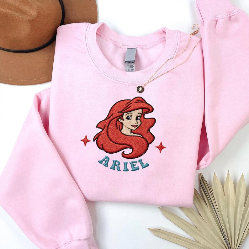 May include: A light pink sweatshirt featuring an embroidered image of Ariel from The Little Mermaid. The design includes Ariel's red hair, face, and the name "ARIEL" in teal lettering. Two red starbursts flank the name. A gold necklace is draped across the sweatshirt.
