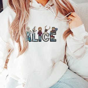 Może przedstawiać: Biała bluza z kapturem z napisem "ALICE" w niebieskiej i czarnej czcionce. Litery ozdobione są postaciami z kresk&oacute;wek. Postacie to m.in. księżniczka, bałwan, dziewczyna, renifer i chłopiec.