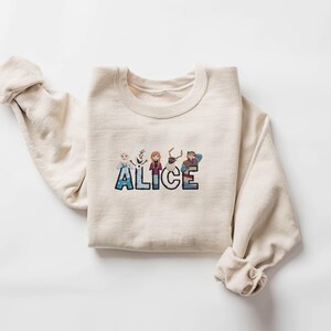 Może przedstawiać: Kremowa bluza z napisem "ALICE" wyhaftowanym kolorową, kresk&oacute;wkową czcionką. Litery ozdobione są postaciami z filmu Kraina Lodu. Rękawy są podwinięte.