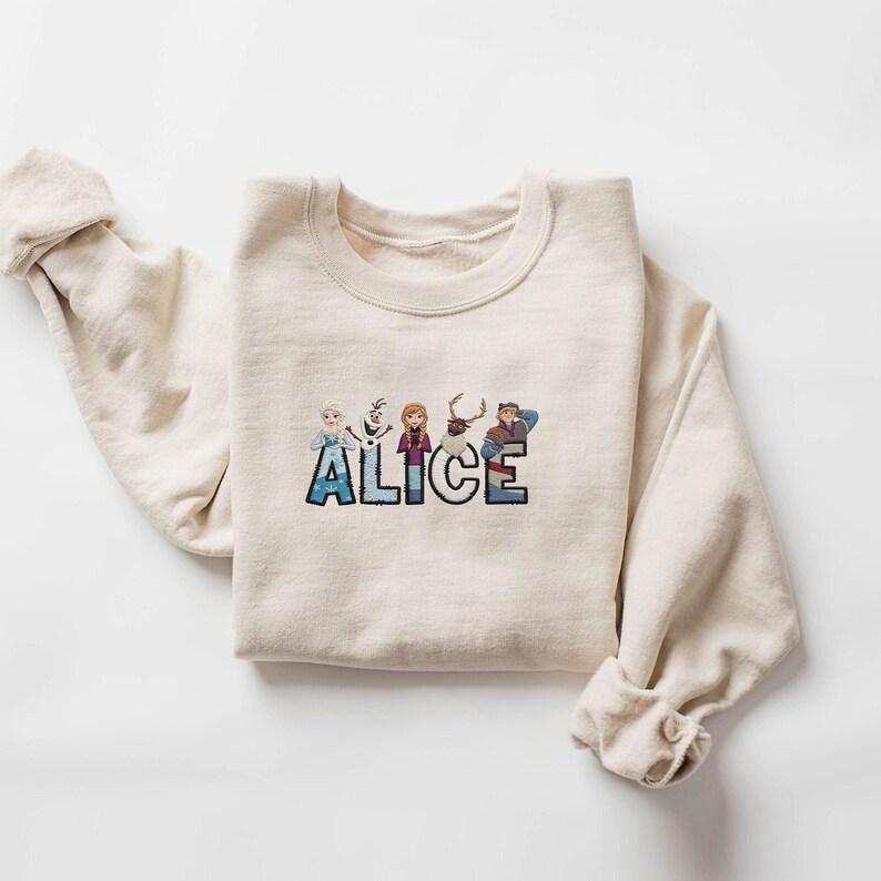 Może przedstawiać: Kremowa bluza z napisem "ALICE" wyhaftowanym kolorową, kresk&oacute;wkową czcionką. Litery ozdobione są postaciami z filmu Kraina Lodu. Rękawy są podwinięte.
