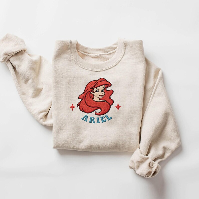 May include: A cream-colored sweatshirt featuring an embroidered image of Ariel from The Little Mermaid. The design includes Ariel's red hair, a blue "ARIEL" text, and red star accents. The sweatshirt is folded with the sleeves rolled up.