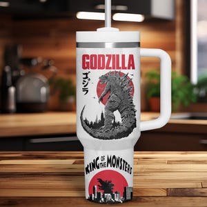 K&ouml;nnte beinhalten: Wei&szlig;er Reisebecher mit Godzilla-Design. Das Design enth&auml;lt das Wort "GODZILLA" in Rot, japanischen Text und eine Schwarz-Wei&szlig;-Illustration von Godzilla. Der Boden des Bechers tr&auml;gt den Text "KING OF THE MONSTERS" in Rot.