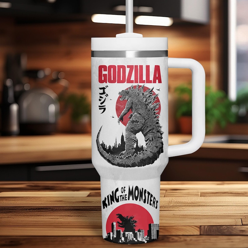 K&ouml;nnte beinhalten: Wei&szlig;er Reisebecher mit Godzilla-Design. Das Design enth&auml;lt das Wort "GODZILLA" in Rot, japanischen Text und eine Schwarz-Wei&szlig;-Illustration von Godzilla. Der Boden des Bechers tr&auml;gt den Text "KING OF THE MONSTERS" in Rot.