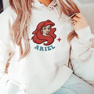 May include: Cream-colored hooded sweatshirt featuring an embroidered image of Ariel with red hair and the name "ARIEL" in blue lettering. Two red starbursts flank the image. The sweatshirt has a drawstring and a kangaroo pocket.