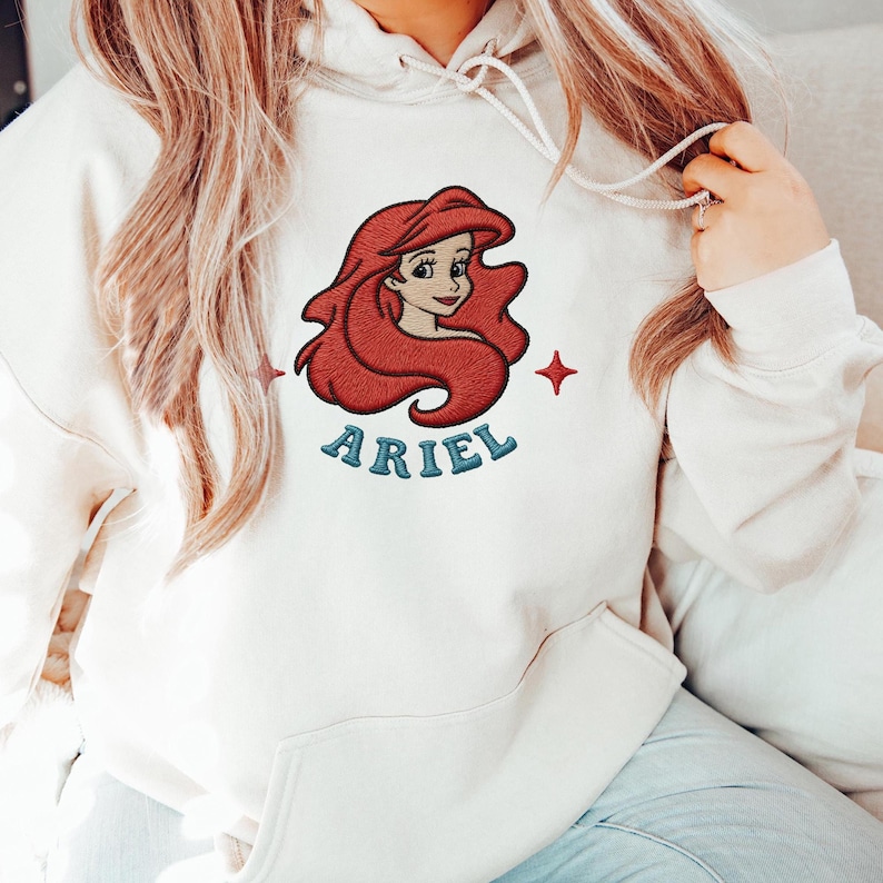 May include: Cream-colored hooded sweatshirt featuring an embroidered image of Ariel with red hair and the name "ARIEL" in blue lettering. Two red starbursts flank the image. The sweatshirt has a drawstring and a kangaroo pocket.