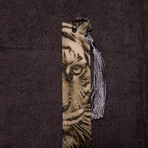 May include: A rectangular bookmark featuring a detailed black and white image of a tiger's face. The bookmark has a silver and black tassel attached to the top. The tiger's eye is the focal point of the design.