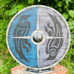 May include: A round wooden shield with a silver metal rim and a central boss. The shield is divided into blue and grey sections, each featuring a stylized black raven design. The shield is mounted on a metal stand.