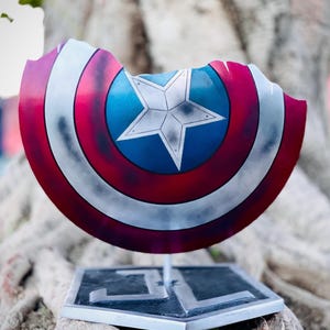 May include: A Captain America shield replica with a distressed design. The shield features a blue central disc with a white star, encircled by red and white stripes. It is mounted on a silver stand with a square base.