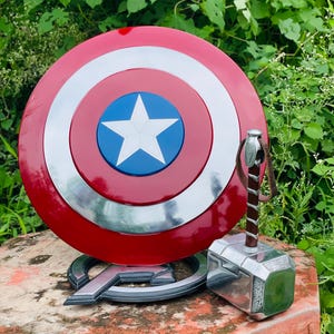 May include: A Captain America shield with a white star on a blue circle, encircled by red and silver rings. Beside it is Thor's hammer, Mjolnir, with a brown leather strap and a silver head. The shield rests on an Avengers logo base.