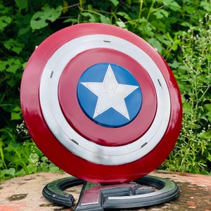 May include: A Captain America shield replica, with a red, white, and blue colour scheme. The shield has a circular design with a white star on a blue centre, surrounded by white and red concentric circles. It rests on a black base with the Avengers logo.