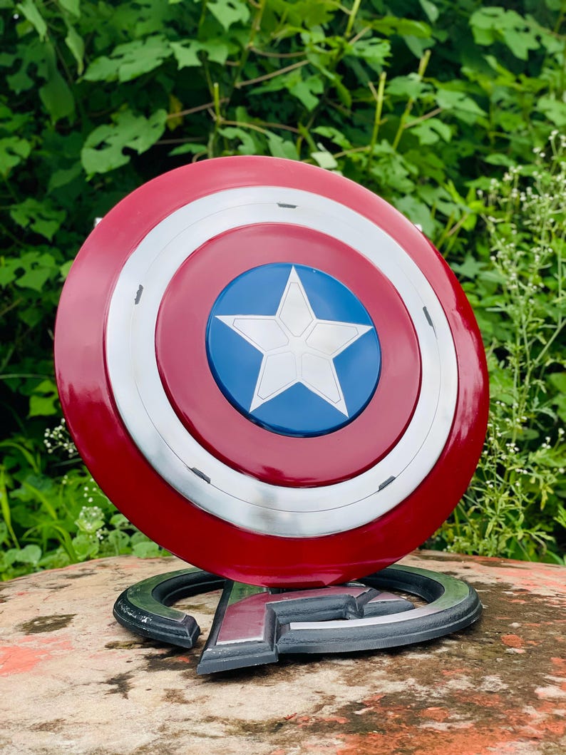 May include: A Captain America shield replica, with a red, white, and blue colour scheme. The shield has a circular design with a white star on a blue centre, surrounded by white and red concentric circles. It rests on a black base with the Avengers logo.