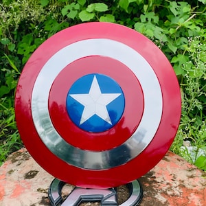 May include: A red, white, and blue Captain America shield with a white star at its centre. The shield is round with a silver border. It is displayed on a stand with the Avengers logo.