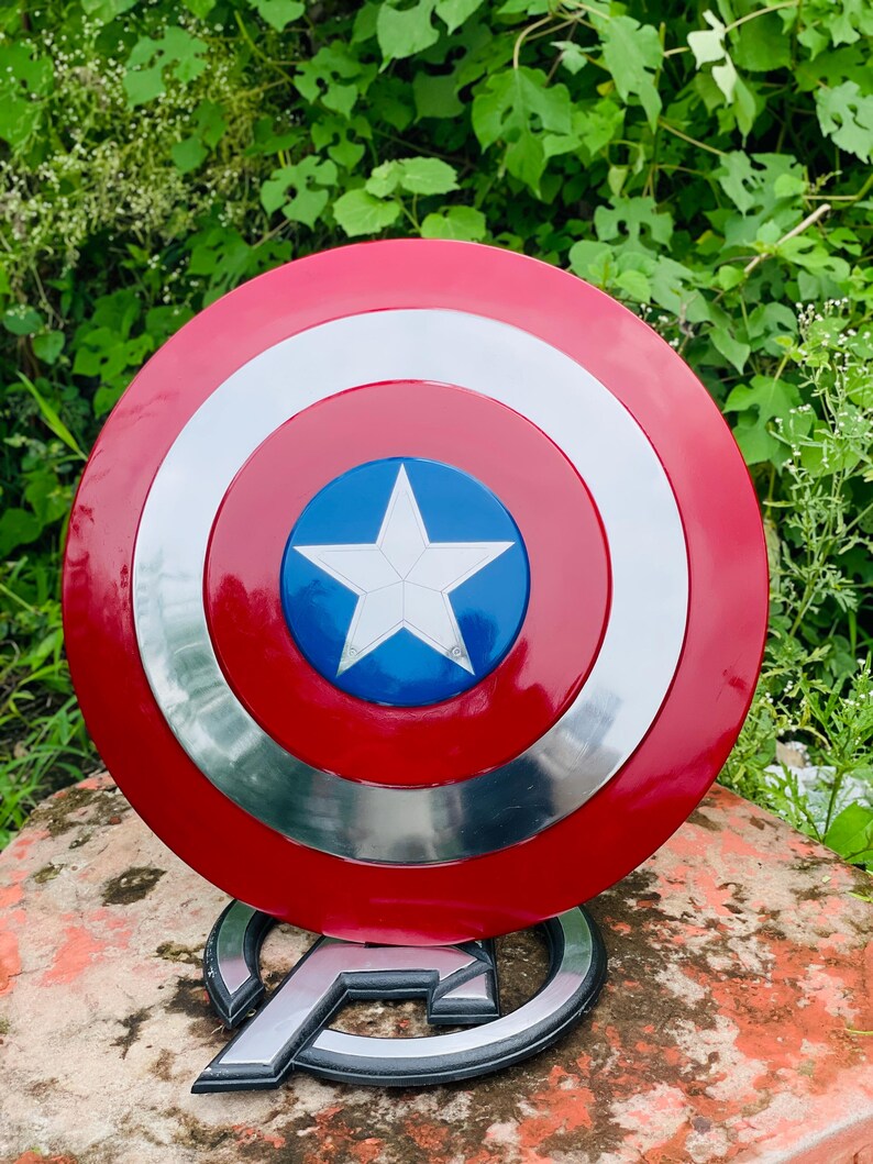 May include: A red, white, and blue Captain America shield with a white star at its centre. The shield is round with a silver border. It is displayed on a stand with the Avengers logo.