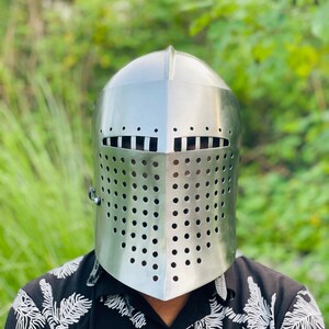 K&ouml;nnte beinhalten: Ein silberner mittelalterlicher Ritterhelm mit einem Visier, das mit zahlreichen kleinen, gleichm&auml;&szlig;ig verteilten L&ouml;chern versehen ist. Der Helm hat eine abgerundete Oberseite und einen vertikalen Grat. Der Hintergrund ist verschwommenes gr&uuml;nes Laub.