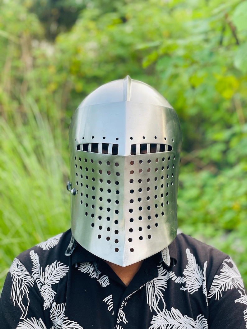 K&ouml;nnte beinhalten: Ein silberner mittelalterlicher Ritterhelm mit einem Visier, das mit zahlreichen kleinen, gleichm&auml;&szlig;ig verteilten L&ouml;chern versehen ist. Der Helm hat eine abgerundete Oberseite und einen vertikalen Grat. Der Hintergrund ist verschwommenes gr&uuml;nes Laub.