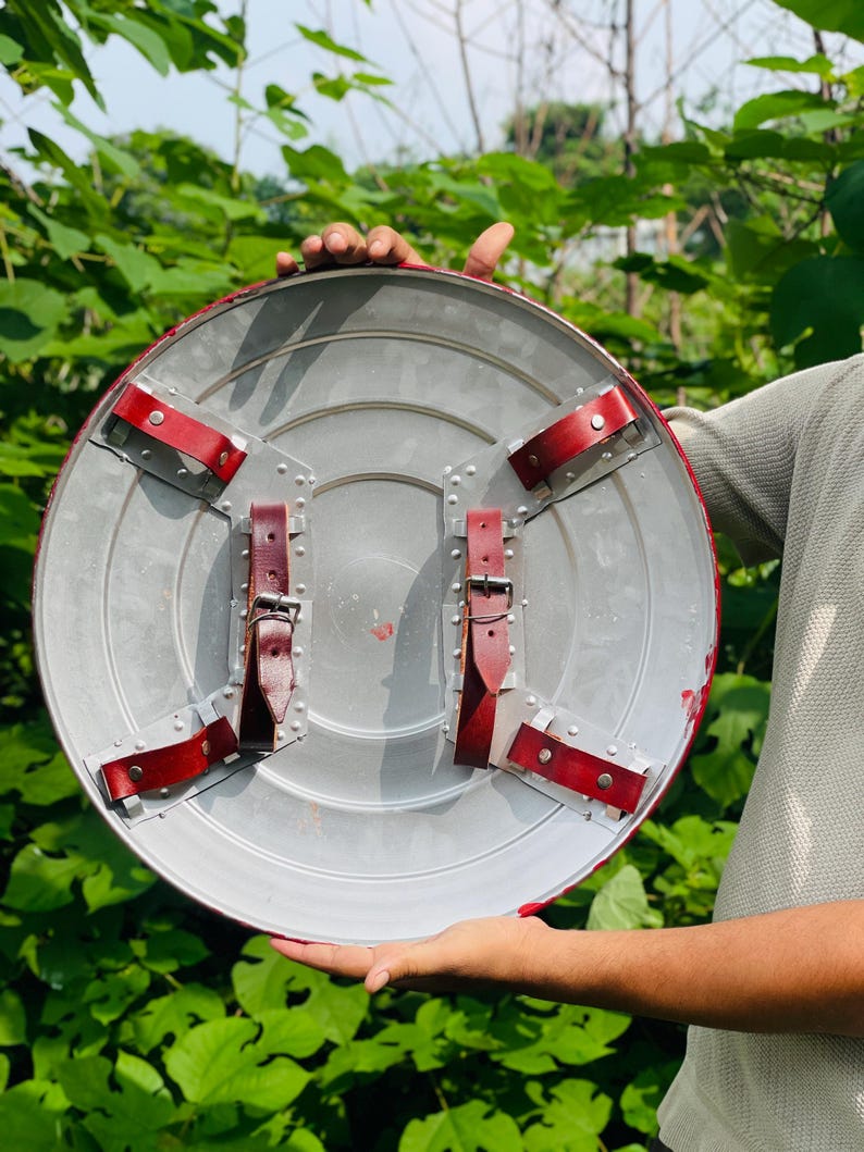 May include: A silver shield with red edging and leather straps. The back of the shield has four red leather straps, secured with metal rivets and buckles. The shield is held against a backdrop of green leaves.