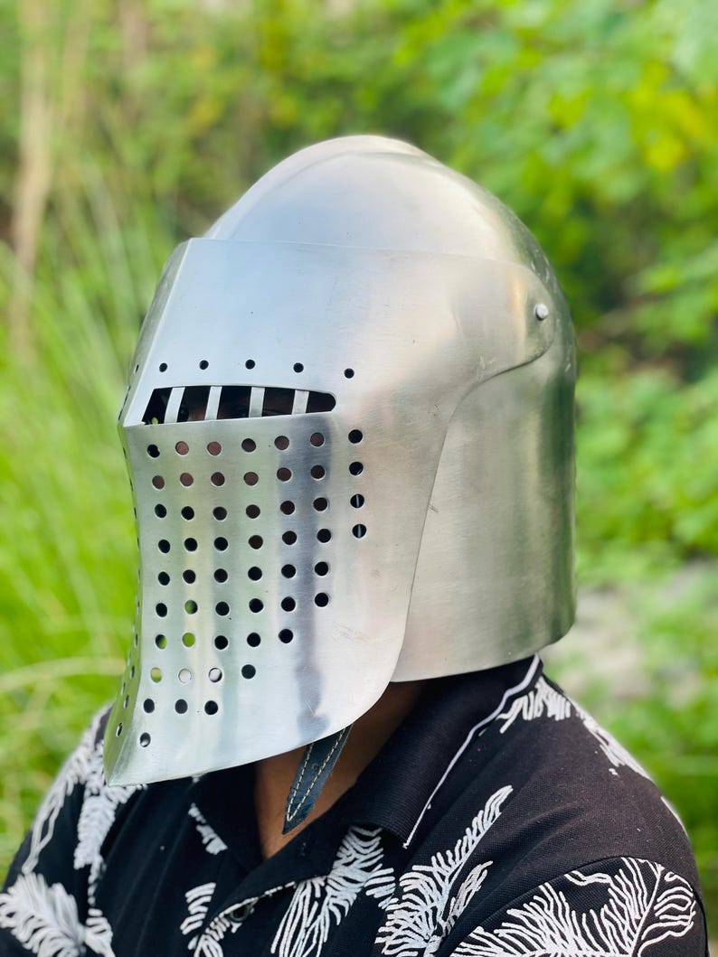 K&ouml;nnte beinhalten: Ein gl&auml;nzender silberner Ritterhelm mit einem Visier, das mit kleinen L&ouml;chern versehen ist. Der Helm hat eine abgerundete Krone und einen Nackenschutz. Der Hintergrund ist ein verschwommenes Gr&uuml;n, was auf eine Umgebung im Freien hindeutet.