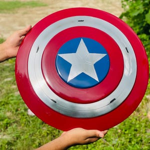 May include: A circular shield in red, white, and blue, featuring a white star on a blue central disc. The design includes a silver band. The shield is held by two hands. The diameter is approximately 76 cm.
