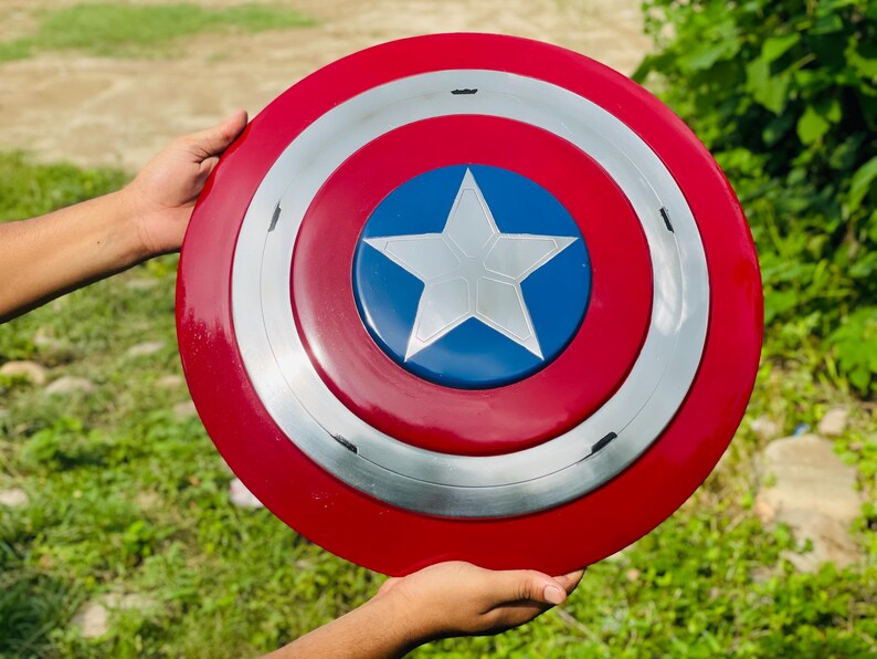 May include: A circular shield in red, white, and blue, featuring a white star on a blue central disc. The design includes a silver band. The shield is held by two hands. The diameter is approximately 76 cm.