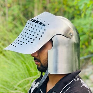 K&ouml;nnte beinhalten: Ein silberfarbener mittelalterlicher Helm mit Visier. Das Visier hat viele kleine, gleichm&auml;&szlig;ig verteilte L&ouml;cher. Der Helm sch&uuml;tzt Kopf und Gesicht, mit gew&ouml;lbter Oberseite und Nackenschutz. Das Visier ragt nach vorne.