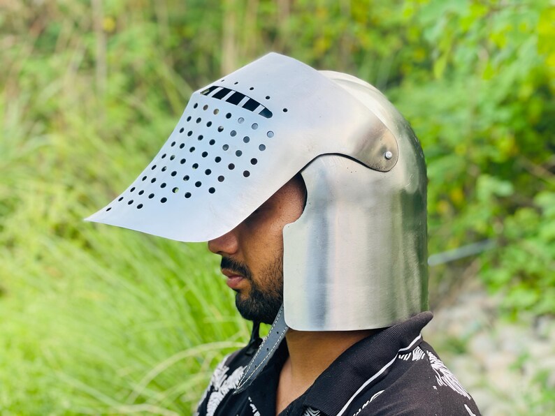 K&ouml;nnte beinhalten: Ein silberfarbener mittelalterlicher Helm mit Visier. Das Visier hat viele kleine, gleichm&auml;&szlig;ig verteilte L&ouml;cher. Der Helm sch&uuml;tzt Kopf und Gesicht, mit gew&ouml;lbter Oberseite und Nackenschutz. Das Visier ragt nach vorne.