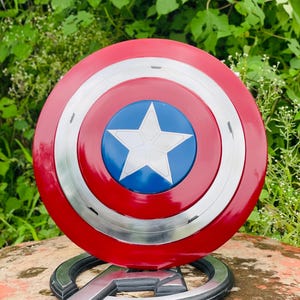 May include: A Captain America shield replica, with a red, white, and blue design. The shield has a central blue circle with a white star, encircled by red and silver rings. It is displayed on a silver Avengers logo base.