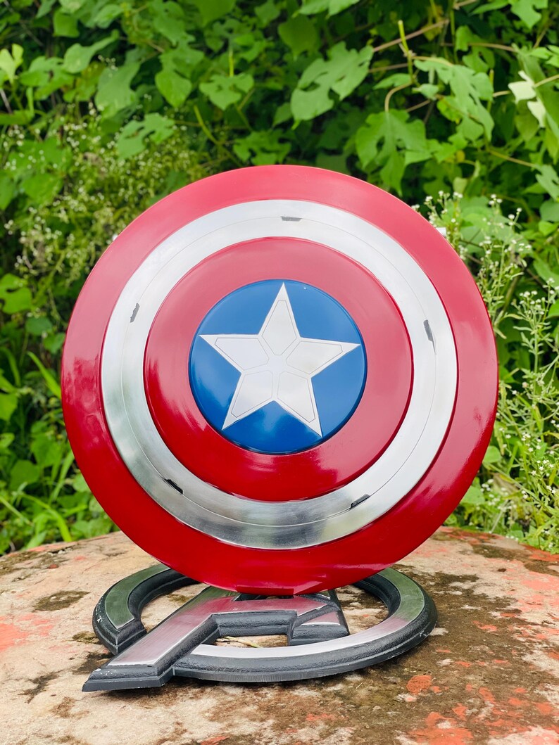 May include: A Captain America shield replica, with a red, white, and blue design. The shield has a central blue circle with a white star, encircled by red and silver rings. It is displayed on a silver Avengers logo base.